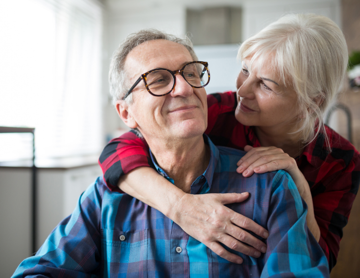 Picture of an older couple, a man in glasses is seated and a woman is lovingly embracing him from behind.