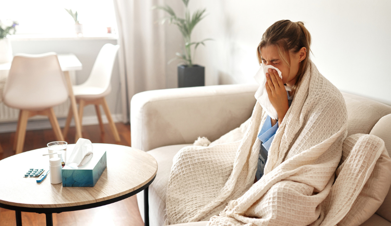 A young woman feeling unwell is wrapped snugly in a blanket while sitting on her couch