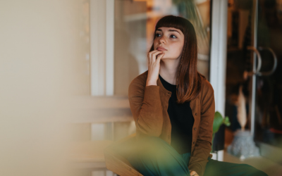  A young woman sits indoors in a thoughtful pose, wearing casual but stylish attire and a pensive expression. 