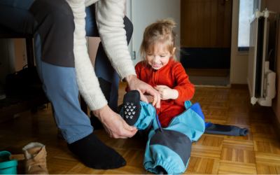Father helping toddler get dressed in overall at hallway, child learning to put on clothes before going outside in cold season. 