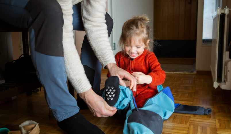 Father helping toddler get dressed in overall at hallway, child learning to put on clothes before going outside in cold season. 