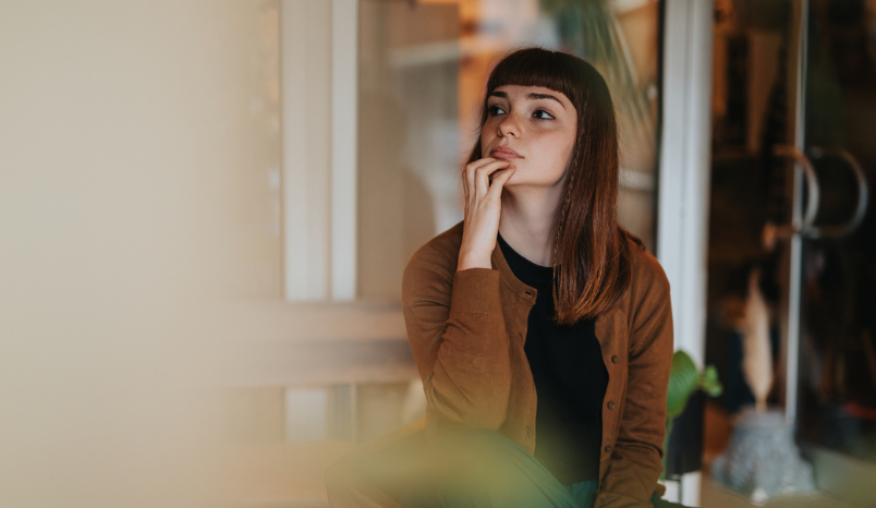 A young woman sits indoors in a thoughtful pose, wearing casual but stylish attire and a pensive expression. 