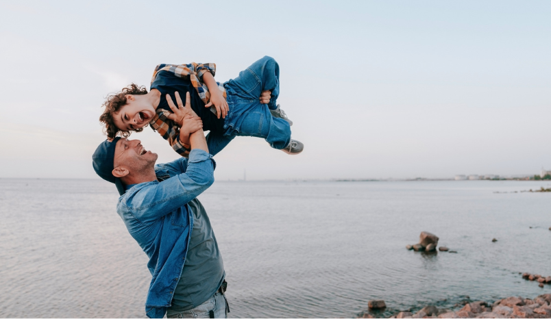 Man on beach playfully lifts young son into the air.