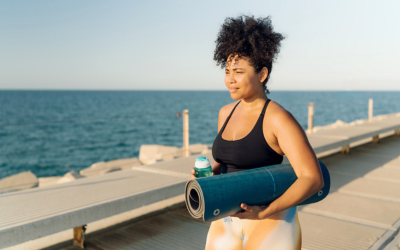 Plus size Hispanic sportswoman holding yoga mat and water bottle walking on pier by the sea 