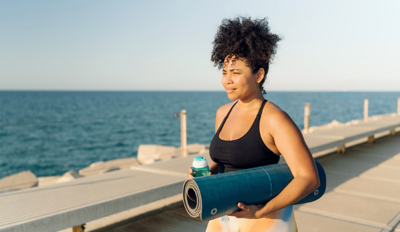  Plus size Hispanic sportswoman holding yoga mat and water bottle walking on pier by the sea 