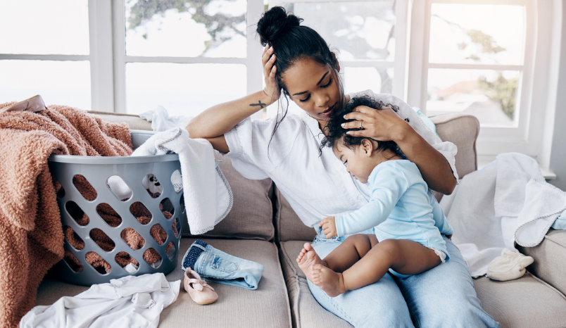 Mom sits on sofa with baby next to full laundry bin.