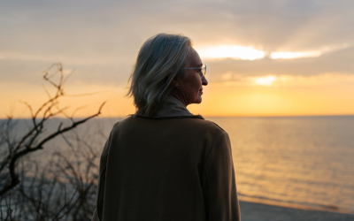  Senior woman wearing glasses and a coat standing on the seashore at sunset.