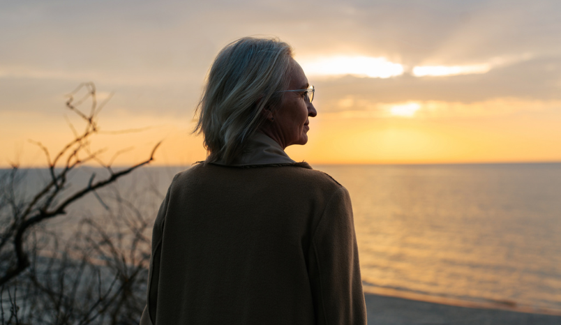 Senior woman wearing glasses and a coat standing on the seashore at sunset.