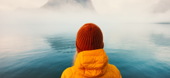 Woman in winter coat looks out onto foggy waters.