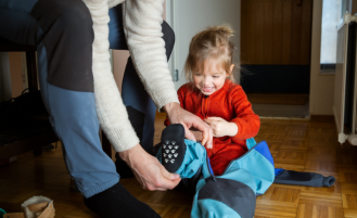 Father helping toddler get dressed in overall at hallway, child learning to put on clothes before going outside in cold season. 