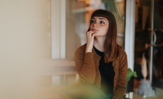 A young woman sits indoors in a thoughtful pose, wearing casual but stylish attire and a pensive expression. 