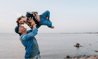 Man on beach playfully lifts young son into the air.
