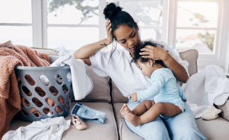 Mom sits on sofa with baby next to full laundry bin.